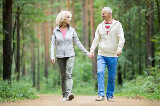 Vanhempi pari aktiivisissa vaatteissa kävelemässä metsätietä pitäen kädestä ja katsoen toisiaan
 - Valokuva, kuva