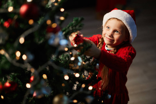 Маленькая девочка висит украшения на ветках елки с искрящимися огнями феи
 - Фото, изображение