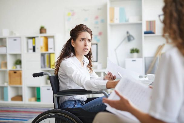 Spöttische Geschäftsfrau im Rollstuhl schaut ihren Kollegen während der Diskussion über Dokumente bei einem Treffen an - Foto, Bild