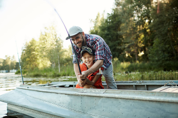 Отец учит своего счастливого сына рыбалке, он помогает ему держать удочку, они сидят на лодке
 - Фото, изображение