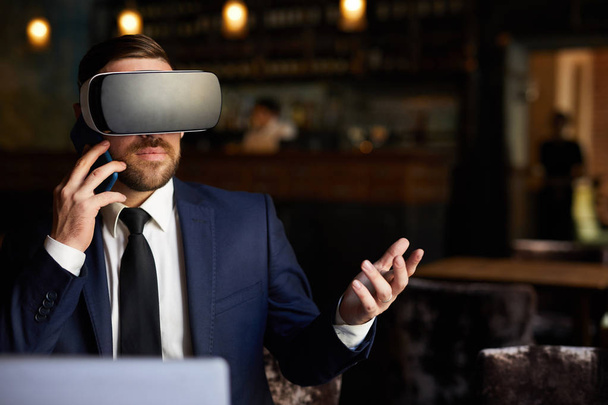 Vakava luottavainen moderni parrakas liikemies virtuaalitodellisuudessa kuulokkeet eleitä puhuessaan matkapuhelin ja katsella video ravintolassa
 - Valokuva, kuva
