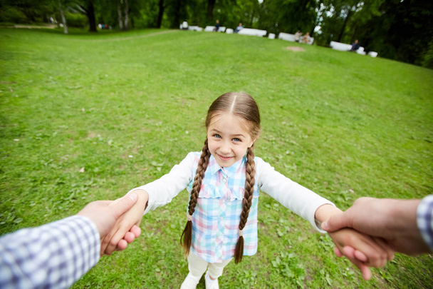 Roztomilá holčička, držící se v rukou svých rodičů, zatímco stojí na zeleném trávníku v parku - Fotografie, Obrázek