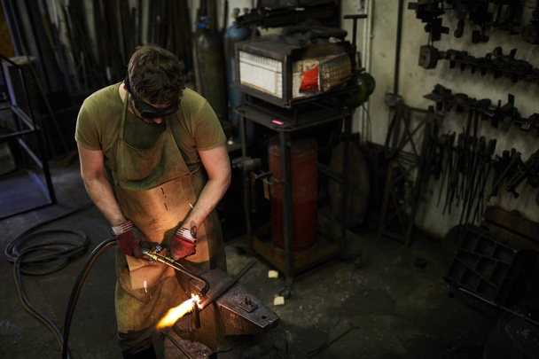 Concentrated busy blacksmith in dark safety goggles moving torch while cutting metal on anvil, he welding in workshop - 写真・画像