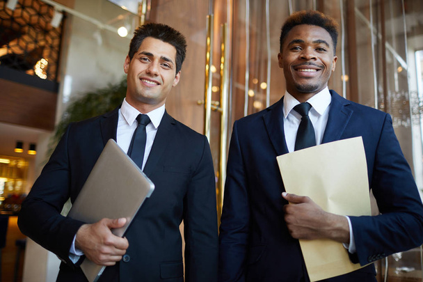 Cheerful confident multi-ethnic business partners in formal suits standing in modern lobby and looking at camera, they holding laptop and papers - Fotó, kép