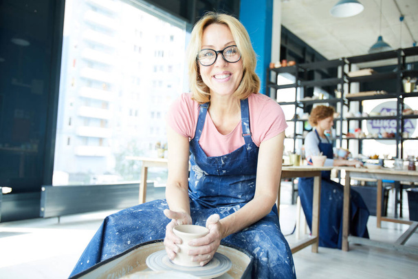 Happy young master of pottery looking at you while siting by pottery-wheel in her workshop - 写真・画像