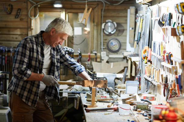 Starszy mistrz obróbki drewna w odzieży roboczej stojącej w warsztacie i piłowaniu drewna - Zdjęcie, obraz