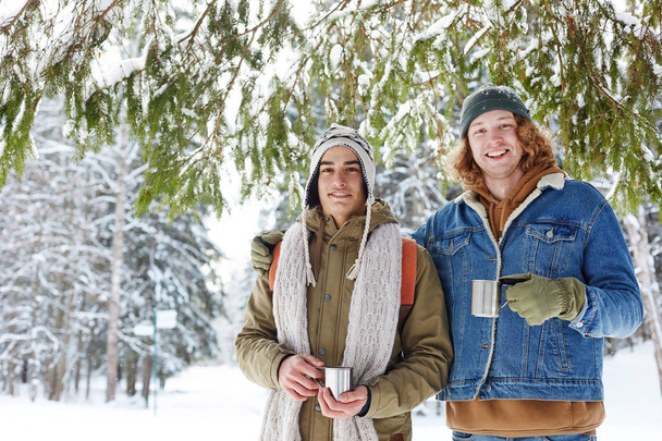 Talia się portret dwóch młodych mężczyzn w ośrodku zimowym stwarzające kubki z gorącym kakao i patrząc na aparat uśmiechnięty, Kopiuj przestrzeń - Zdjęcie, obraz
