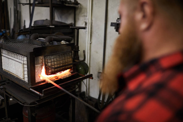 Ferreiro moderno em pé pelo forno em chamas e mantendo a extremidade do pau de ferro quente dentro dele
 - Foto, Imagem