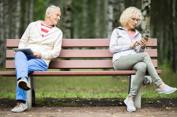 Старший чоловік з тачпадкою дивиться на жіночий смс в смартфоні на іншій стороні лави
 - Фото, зображення