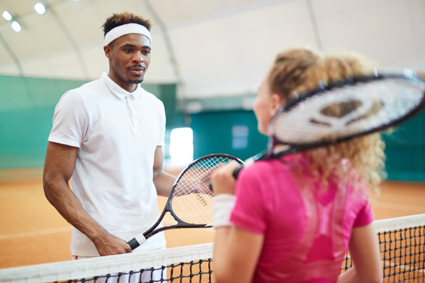 Joven tenista afroamericano hablando con su compañero de juego detrás de la red en la cancha
 - Foto, imagen