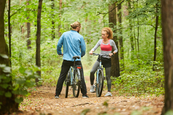 Menina ativa e cara com bicicletas em pé no caminho da floresta e discutindo que maneira de escolher para o passeio
 - Foto, Imagem
