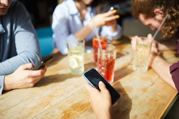A fiatalok okostelefonjaikat használják, miközben a fa asztalnál ülnek a napsütéses napon és iszogatnak. - Fotó, kép