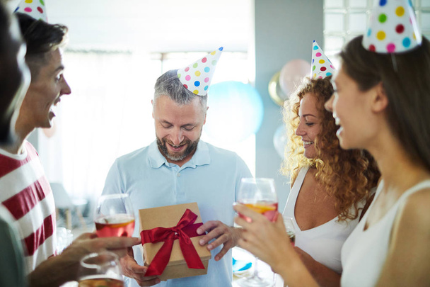Щасливий чоловік відкриває коробку з подарунком на день народження від своїх друзів під час тостів для нього
 - Фото, зображення