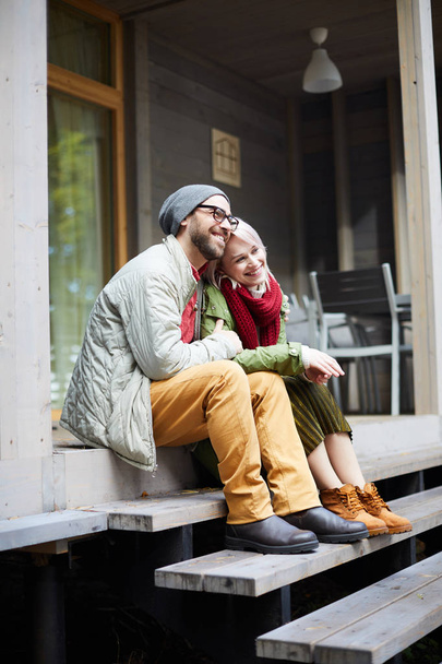 Young attractive fashionable Caucasian couple sitting on wooden steps of new house, embracing each other and smiling cheerfully - Foto, immagini