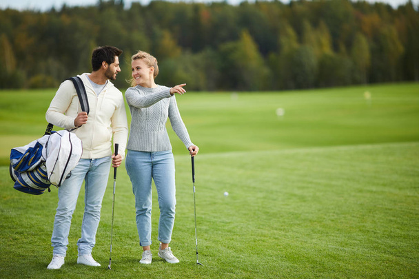 Young woman showing her boyfriend nice place for playing golf while both standing on large green field - Foto, afbeelding