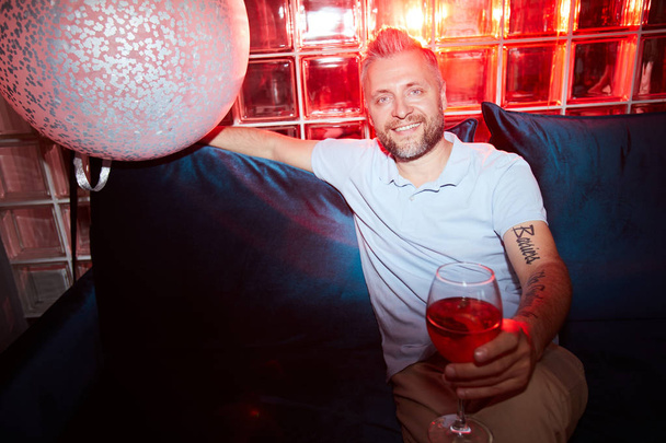 Uomo felice con bevanda fatta in casa in bicchiere di vino che si trova sul divano a casa festa e ti guarda
 - Foto, immagini