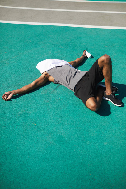 Неузнаваемый измученный молодой черный мужчина с полотенцем на лице лежит на стадионе и держит бутылку воды на открытом воздухе
 - Фото, изображение