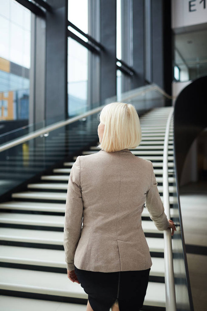 Back view of elegant blond businesswoman ascending by staircase along large window - Foto, immagini