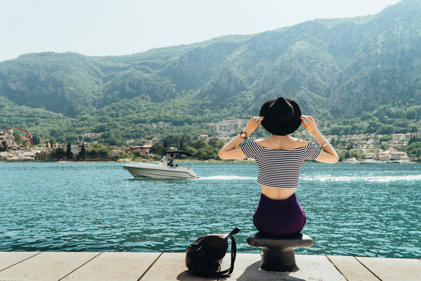 Κορίτσι ταξιδιώτης σε ένα μαύρο καπέλο και ένα ριγέ μπλουζάκι κάθεται στην προβλήτα στον κόλπο του azure που περιβάλλεται από βουνά. Δίπλα της είναι το σακίδιο. - Φωτογραφία, εικόνα