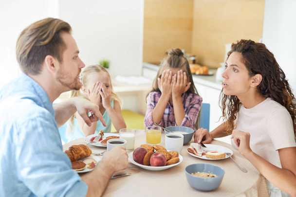 Молода роздратована пара має суперечки на сніданок, а їх дочки сидять поруч і покривають обличчя руками
 - Фото, зображення