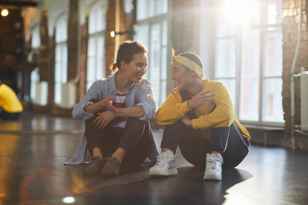 zwei fröhliche freundliche Mädchen, die nach dem Training auf dem Boden sitzen und ein paar Trainingsutensilien besprechen - Foto, Bild