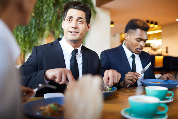 positiver gutaussehender junger Geschäftsmann im formellen Anzug, der am Tisch sitzt und Salat isst, während er mit Kollegen beim Abendessen im Restaurant plaudert - Foto, Bild