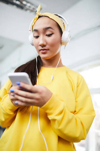 Menina ativa em camisola amarela e headband ouvir música em fones de ouvido ao escolher a trilha sonora da lista de reprodução
 - Foto, Imagem