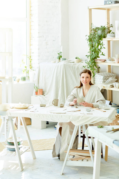 Щаслива молода жінка в робочому одязі, сидячи за столом у своїй майстерні, дивлячись на камеру і роблячи посуд з глини
 - Фото, зображення
