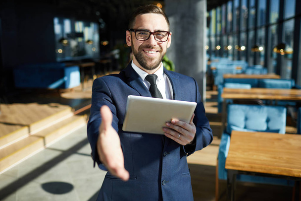 Επιχειρηματίας με μεγάλο χαμόγελο κρατώντας Touchpad στο ένα χέρι, ενώ δίνοντας το άλλο σε πελάτη του καφέ για χειραψία - Φωτογραφία, εικόνα