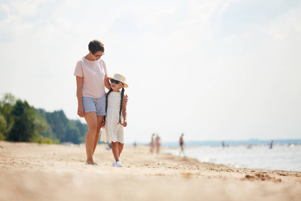 Full length portrait of loving modern mom embracing daughter while enjoying walk along beach during Summer vacation, copy space - Фото, изображение