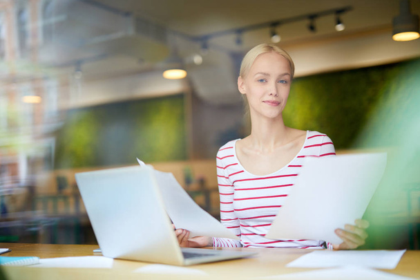Młoda blondynka bizneswoman patrząc na ciebie podczas pracy z papierami przed laptopem - Zdjęcie, obraz
