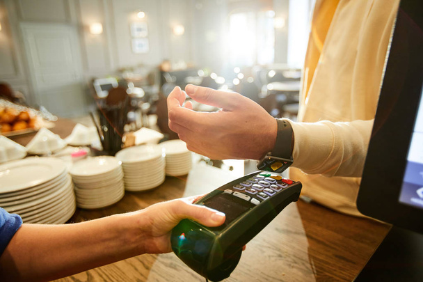 Kassiererin hält Zahlungsgerät, während einer der Kunden mit Smartwatch sein Handgelenk darüber hält - Foto, Bild