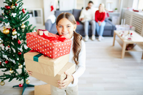 Веселая маленькая девочка со стопкой подарков несёт их в свою комнату, чтобы распаковать
 - Фото, изображение
