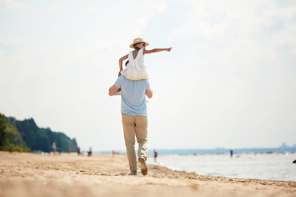 Vista trasera del joven sosteniendo a su pequeña hija en hombros mientras camina por la playa de arena el fin de semana
 - Foto, imagen