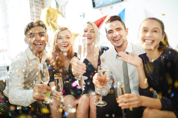 Jonge vrolijke vrienden met fluiten van champagne plezier hebben in confetti regen op verjaardagsfeestje - Foto, afbeelding