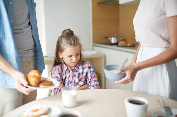 Naughty little girl expressing dislike while her parents offering her homemade pastry and muesli for breakfast - Фото, зображення