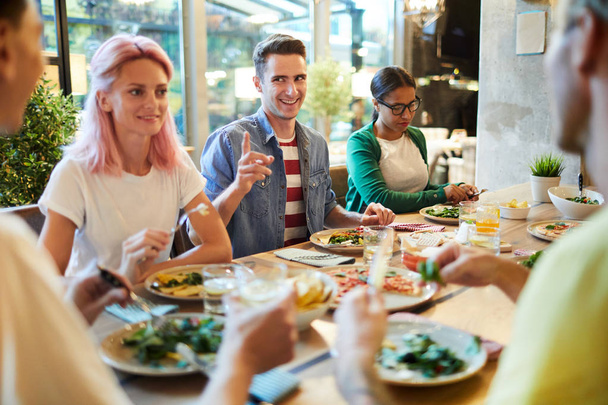 Счастливый парень указывает на одного из своих друзей за ужином в кафе, разговаривая с ней
 - Фото, изображение
