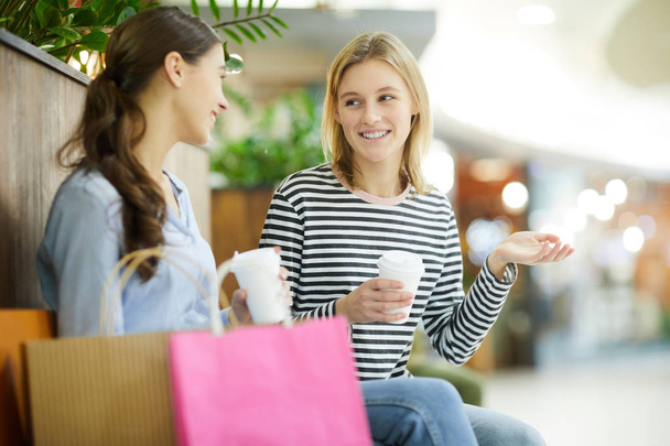 Дві дружні випадкові дівчата з напоями розмовляють під час відпочинку в сучасному торговому центрі після покупок
 - Фото, зображення