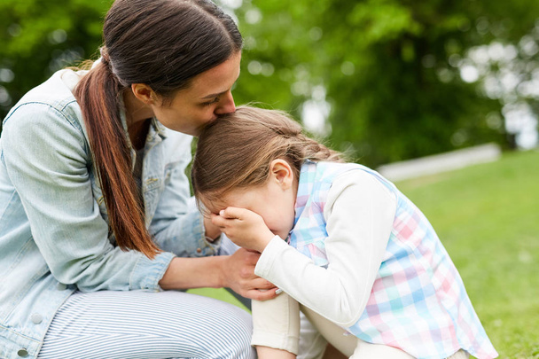 Young woman kissing her crying daughter on head, embracing and comforting her during chill in park - Фото, зображення