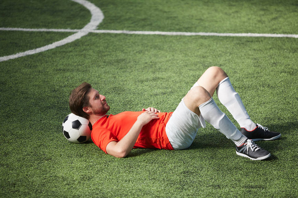 Νέος ποδοσφαιριστής ξεκούραστο ξαπλωμένο στο πράσινο πεδίο με μπάλα ποδοσφαίρου, πίσω από το λαιμό του - Φωτογραφία, εικόνα