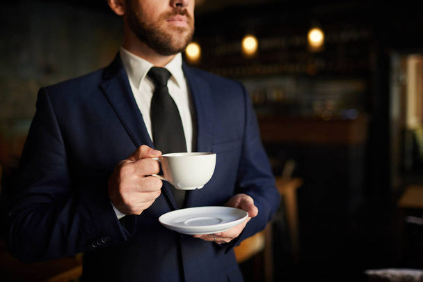 Primer plano de empresario pensativo serio en traje formal de pie y sosteniendo plato mientras bebe café durante el descanso
 - Foto, imagen