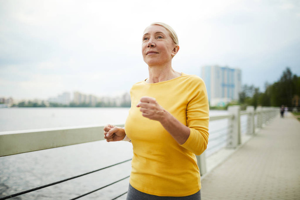 reife, aktive blonde Frau im gelben Pullover joggt morgens auf der Stadtbrücke am Flussufer - Foto, Bild