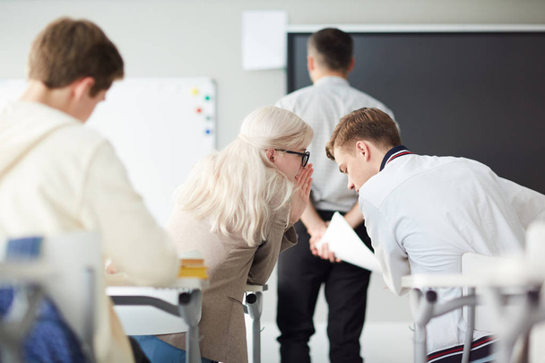 Jóvenes estudiantes chismorreando o discutiendo la respuesta correcta detrás de su profesor en la lección
 - Foto, imagen