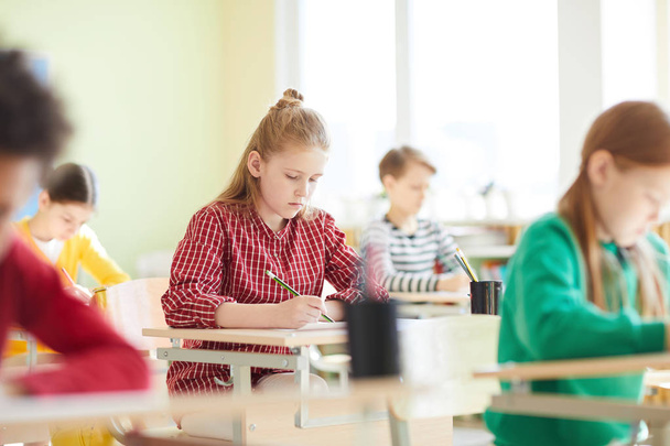 Niños de la escuela llenando el examen en blanco en el examen: colegiala concentrada seria en camisa casual sentada en la mesa y resolviendo la tarea
 - Foto, imagen