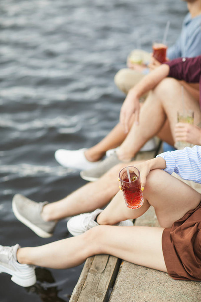 Piernas de cuatro jóvenes amigos sentados junto al agua, disfrutando del día de verano y bebiendo
 - Foto, imagen