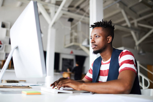 Jonge serieuze zakenman concentreren op het netwerk, terwijl zitten door bureau in de voorkant van computer monitor - Foto, afbeelding