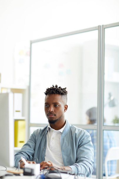 Молодой афро-американский хипстер сидит на рабочем месте перед монитором компьютера и изучает новый язык кода
 - Фото, изображение
