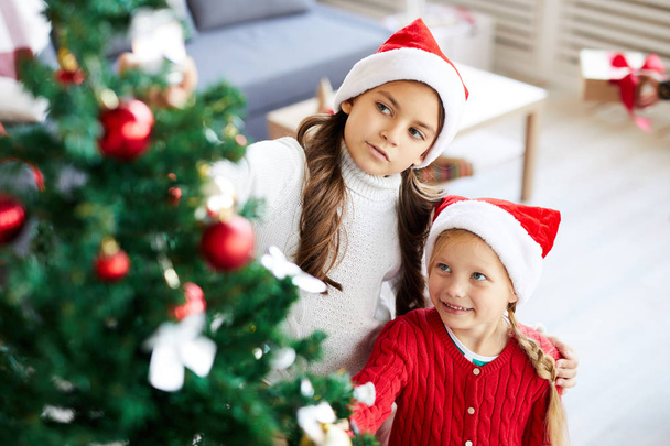 zwei kleine Mädchen in Weihnachtsmannmützen betrachten die Dekoration am Weihnachtsbaum, während sie in der Nähe stehen - Foto, Bild