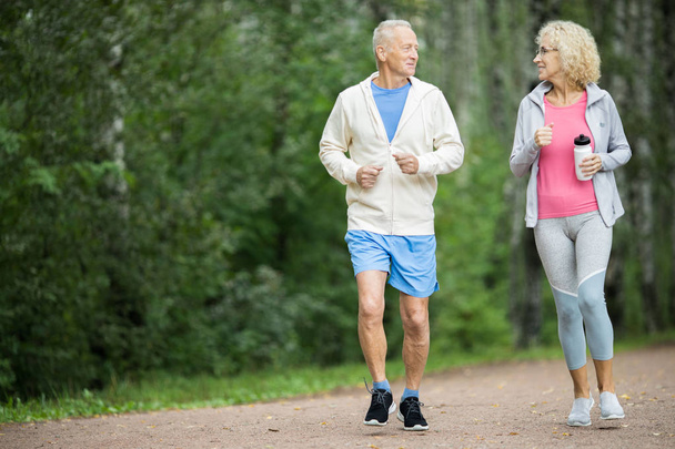 Glückliches rühriges Seniorenpaar joggt morgens im Park unter grünen Bäumen - Foto, Bild