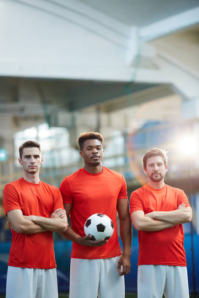 Τρεις νέους ποδοσφαιριστές σε κόκκινα μπλουζάκια και άσπρο σορτσάκι στέκεται μπροστά από την κάμερα στο γήπεδο - Φωτογραφία, εικόνα